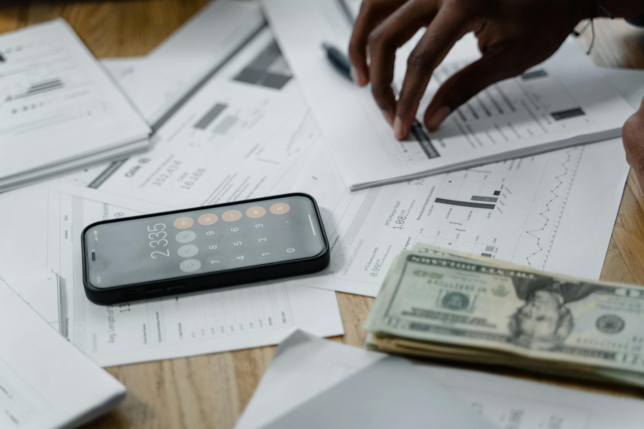 Close-up of hands calculating financial data with reports, smartphone, and cash on table.